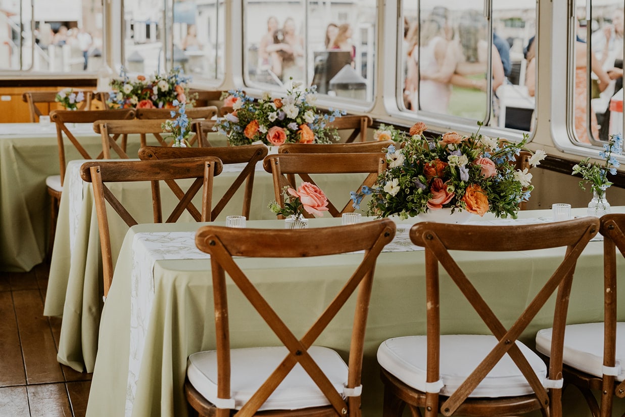 A dockside event boat set for a reception with long tables in sage green linen, wooden cross-back chairs and colorful garden flower centerpieces