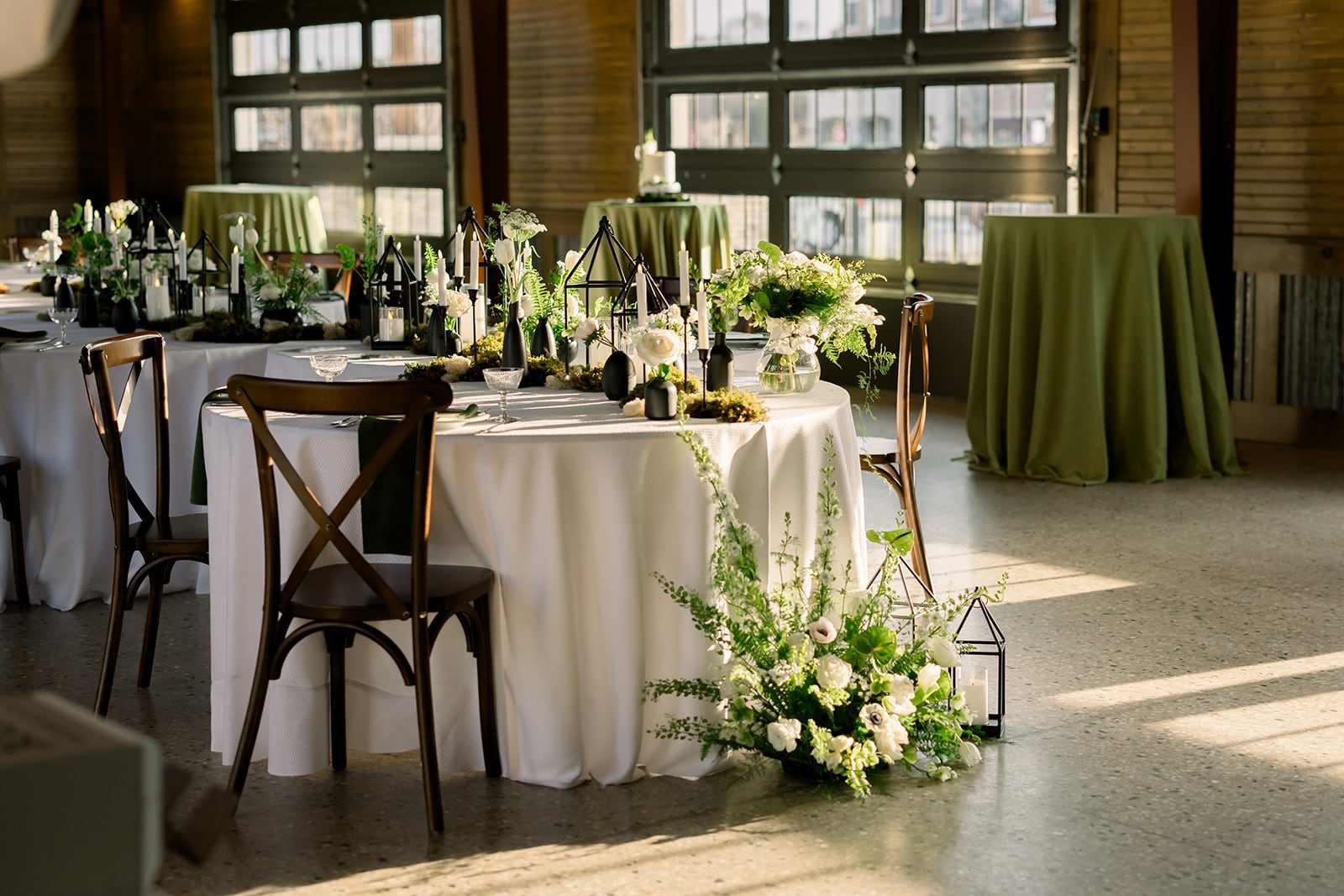 A round table with a white cloth, dark cross-back chairs and a low garden centerpiece of white flowers, moss and black lanterns in a sunlit loft with olive green linen tables